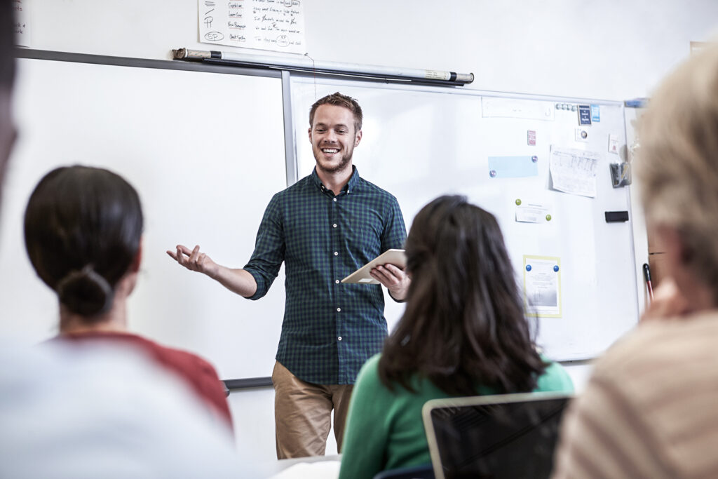 Uma imagem contendo um homem, três pessoas, um quadro branco ao fundo para aula de inglês para iniciantes